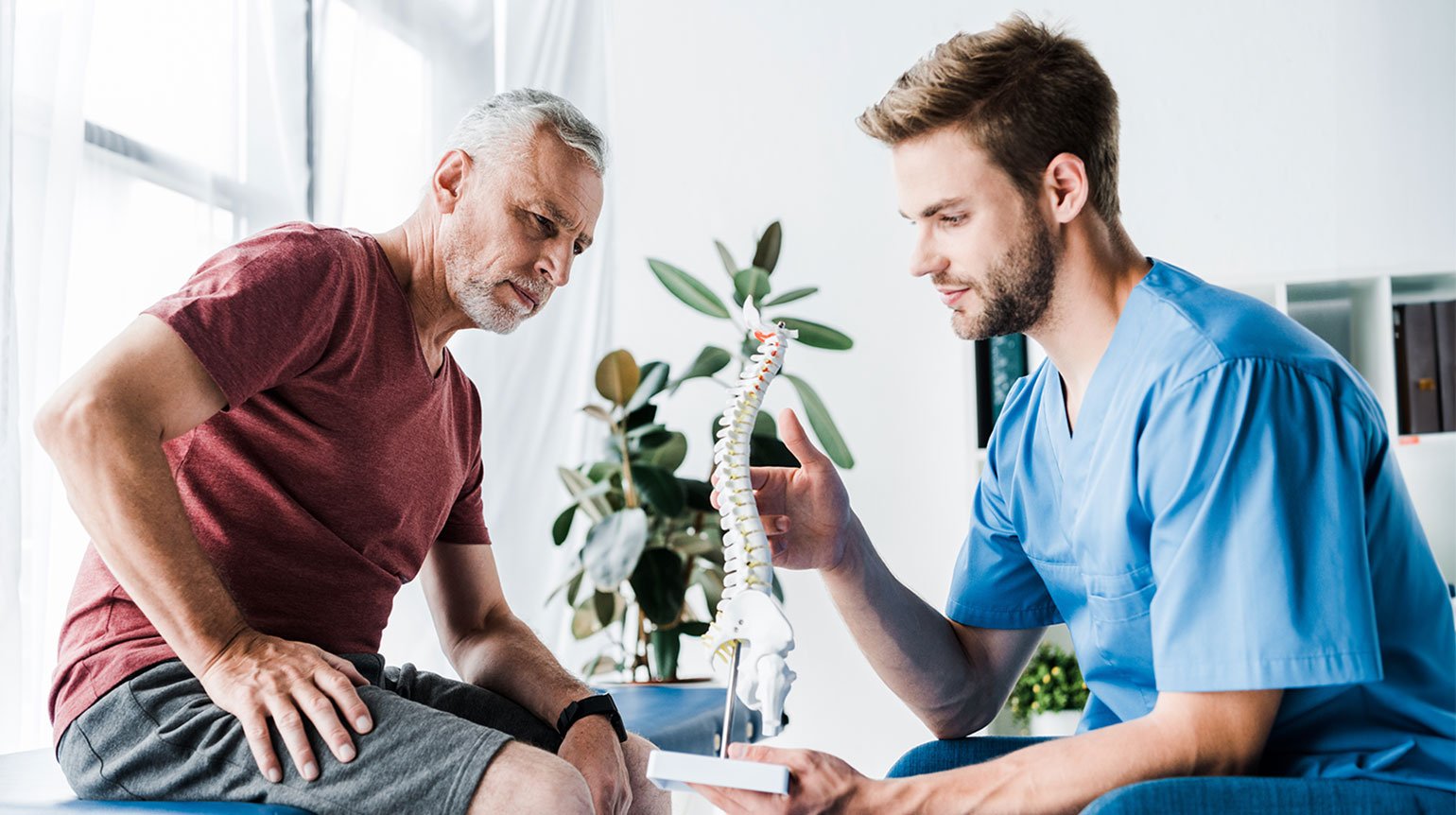 Arzt/Physiotherapeut zeigt Patienten ein Modell der Wirbelsäule. Die Finger seiner rechten Hand berühren die Brustwirbelsäule, vielleicht um einen Prolaps zu besprechen. Beide sitzen sich in einer Praxis gegenüber und betrachten das Modell.