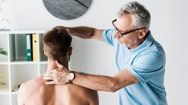 Patient sitzt mit nacktem Oberkörper links im Bild, Rücken uns zugewandt. Arzt/Physiotherapeut steht rechts neben ihm. Er legt die linke Hand an den Nacken und die rechte Hand auf den Kopf des Patienten und betrachtet die Halswirbelsäule.