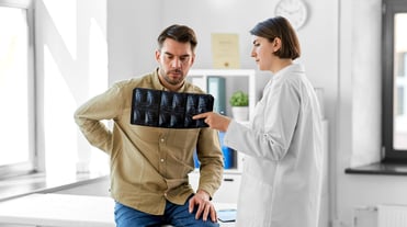Ärztin erklärt Patienten Röntgenbilder in einer Praxis. Er schaut auf die Bilder und hält seinen schmerzenden Rücken. Auf den Bildern ist eine Lendenwirbelsäule zu erkennen. Vermutlich geht es um einen Bandscheibenvorfall.