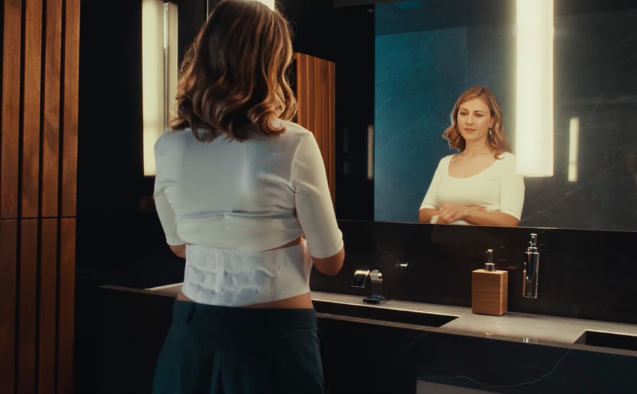 Eine Frau trägt ein ThermaCare Wärmeumschlag auf dem unteren Rücken und schaut in den Spiegel.