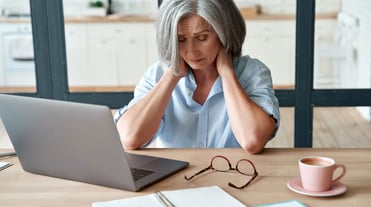 Ein Frau an einem Tisch sitzend, umfasst mit beiden Händen ihren Nackenbereich