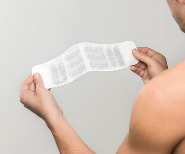Mann mit freiem Oberkörper hält ThermaCare® Wärmeauflage für Schulter und Nacken in beiden Händen