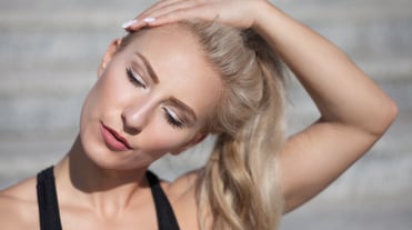 Eine blonde Frau mit Zopf nimmt den Arm über den Kopf, fässt sich an die Schläfe und dehnt die Nackenmuskulatur