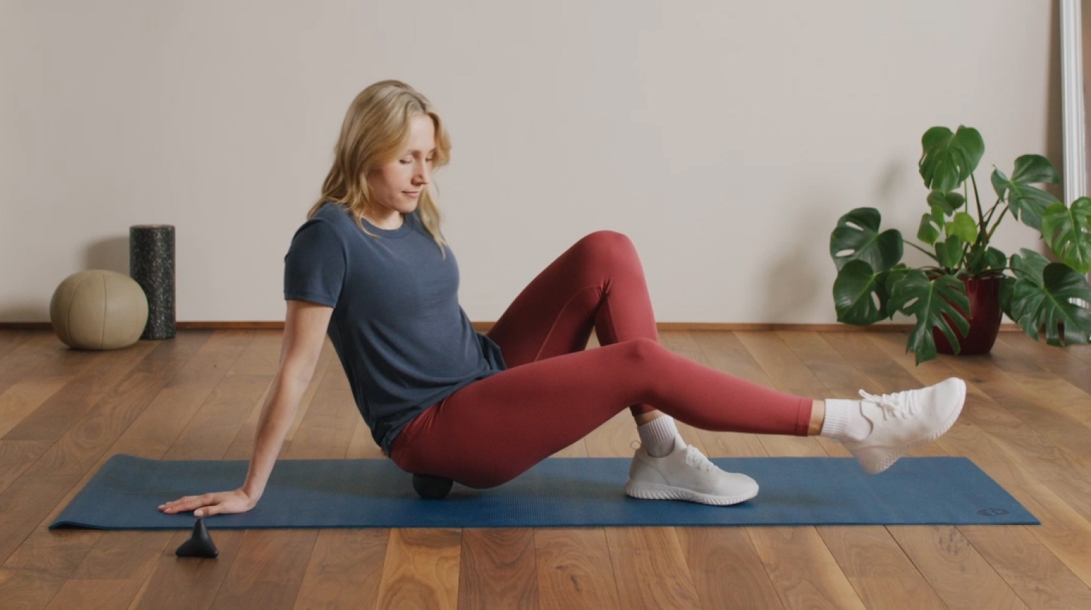 Eine Frau sitzt auf einer Sportmatte und rollt unter ihrem Gesäß eine Ball, um eine Verspannung zu lösen