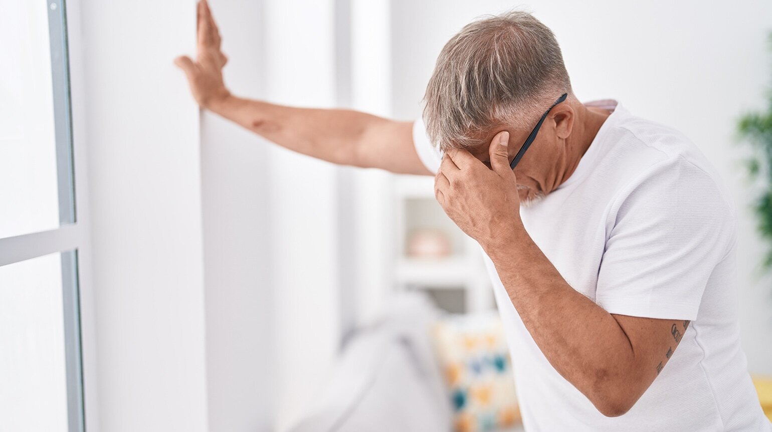 Mann mit weißem Shirt stützt sich mit der rechten Hand an Wand ab, hält sich mit der linken die Schläfe, häufige Haltung bei Schwindel durch die HWS Mann mit weißem Shirt stützt sich mit der rechten Hand an Wand ab, hält sich mit der linken die Schläfe, häufige Haltung bei Schwindel durch die HWS