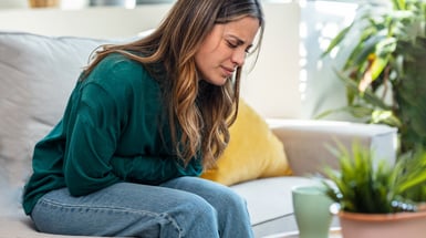 Frau sitzt vornübergebeugt auf einem Bett, hinter sich Kissen. Sie hält sich mit einem schmerzverzerrten Gesicht mit beiden Händen den Bauch. Sie trägt ein weißes Unterhemd und eine blau gestreifte kurze Hose, Bettdecke weiß, Laken blauweiß. 