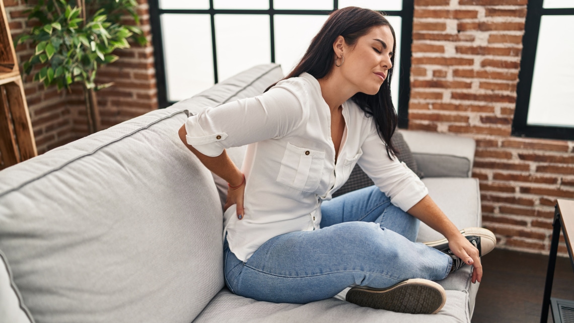 Eine dunkelhaarige Frau sitzt im Schneidersitz auf einer Couch und faesst sich vor Schmerzen an den Ruecken