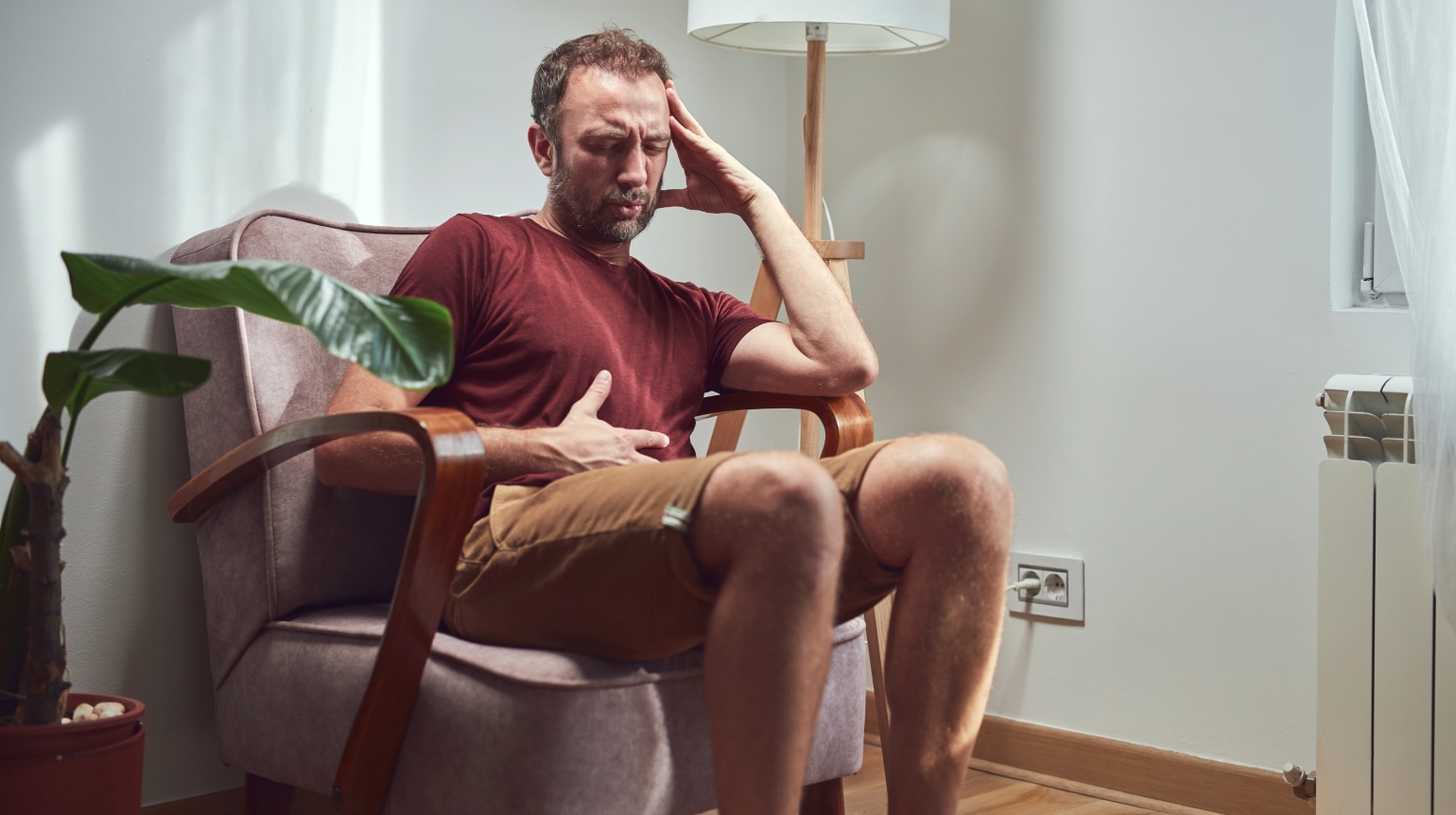 Ein Mann sitzt in einem Sessel und faesst sich vor Schmerzen mit einer Hand an den Kopf, mit der anderen an den Bauch