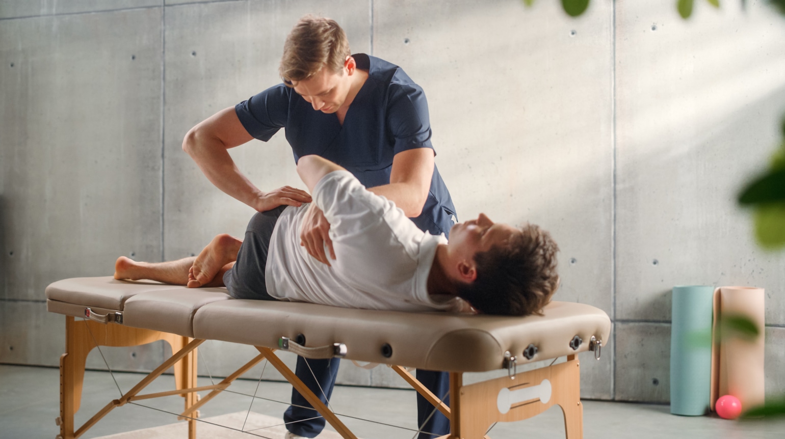 Ein männlicher Physiotherapeut löst eine Blockade im Ruecken eines auch maennlichen Patienten, welcher auf einer Liege liegt