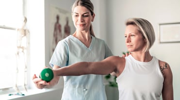 Vorderansicht von zwei Frauen, links Physiotherapeutin, rechts Patientin. Patientin streckt rechten Arm aus, hält kleine grüne Hantel in der Hand. Therapeutin korrigiert Haltung. Im Hintergrund Behandlungszimmer mit Anatomieposter und Fenster.