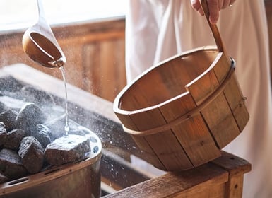 Eine Person mit umgebundenen Handtuch kippt Wasser auf heiße Steine in einer Sauna