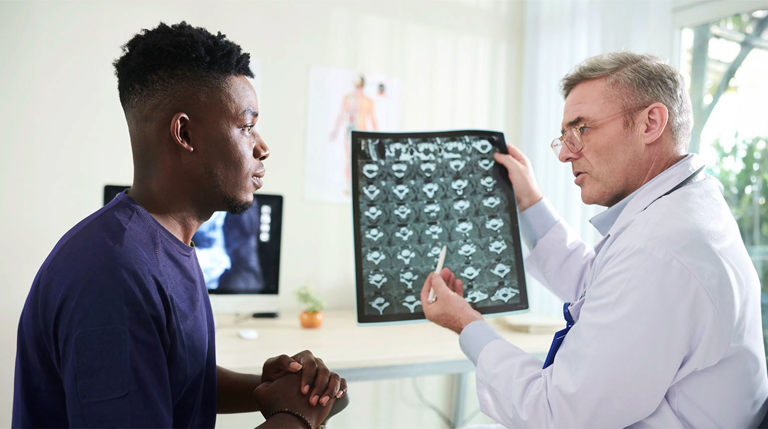 Arzt erklärt Patienten MRT-Bilder eines Bandscheibenvorfalls in der Brustwirbelsäule. Er deutet mit Kugelschreiber auf eine Stelle im Bild. Beide sehen sich an, Patient hört aufmerksam zu. Im Hintergrund Schreibtisch, Bildschirm, kleine Pflanze.