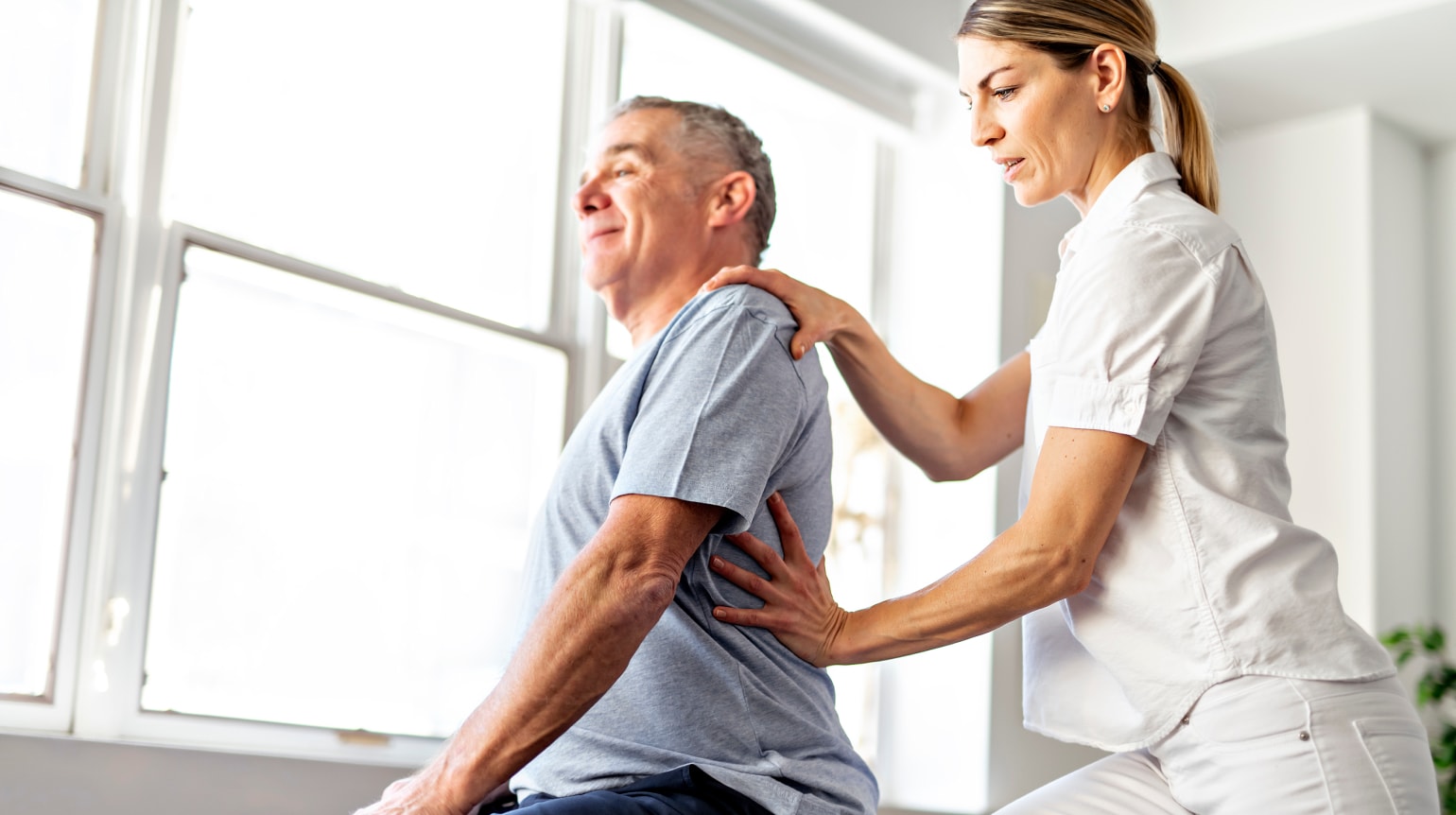 Eine Physiotherapeutin unterstützt einen älteren Mann bei einer Rückenübung in einer Praxis mit großen Fenstern und natürlichem Lichteinfall.