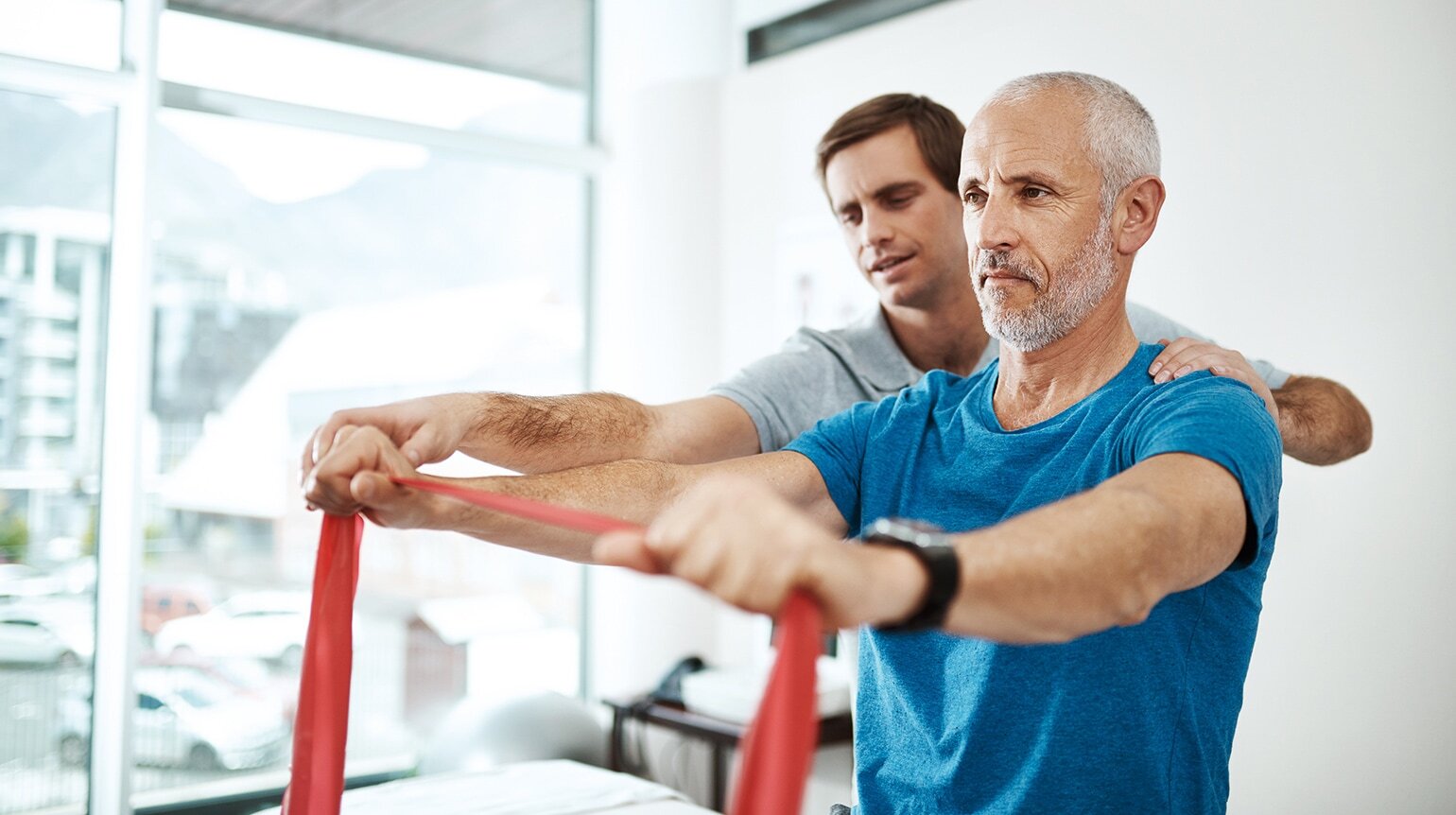 Halbseitenansicht zweier Männer, Patient rechts hält rotes Fitnessband in beiden Händen, Physiotherapeut links dahinter korrigiert Position. Im Hintergrund Behandlungsraum und großes Fenster auf Parkplatz. Halbseitenansicht zweier Männer, Patient rechts hält rotes Fitnessband in beiden Händen, Physiotherapeut links dahinter korrigiert Position. Im Hintergrund Behandlungsraum und großes Fenster auf Parkplatz.