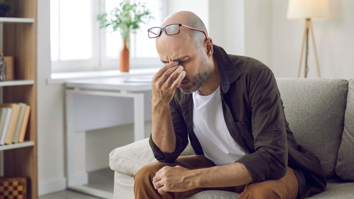 Ein Mann aus einer Couch sitzend fasst mit seiner Hand an seinen Nasenrücken