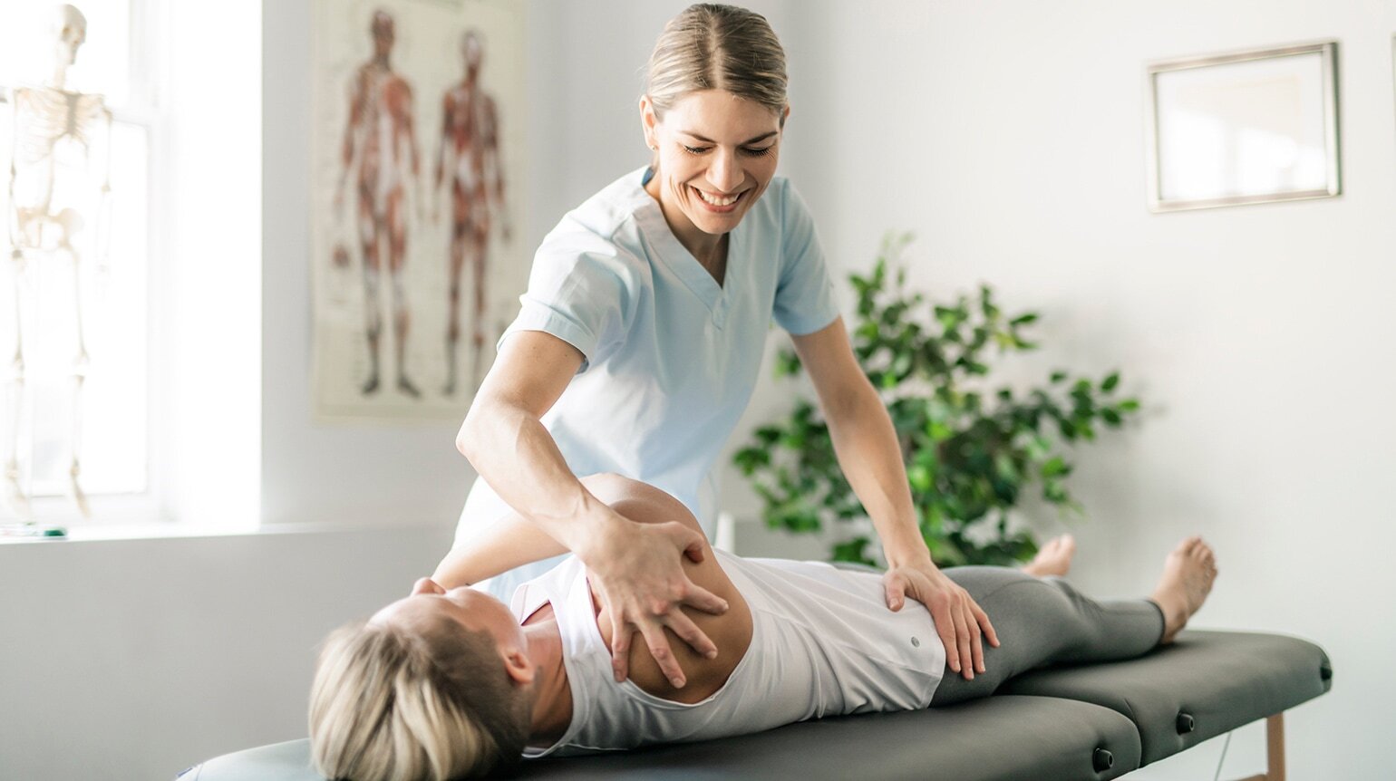 Eine Physiotherapeutin steht an einer Behandlungsliege und führt eine manuelle Therapie an einer auf dem Rücken liegenden Frau durch Eine Physiotherapeutin steht an einer Behandlungsliege und führt eine manuelle Therapie an einer auf dem Rücken liegenden Frau durch
