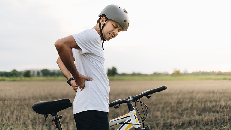 Ein Mann steht mit seinem Fahrrad auf einem Feld und greift sich mit seinem schmerzverzerrtem Gesicht  an den Ruecken