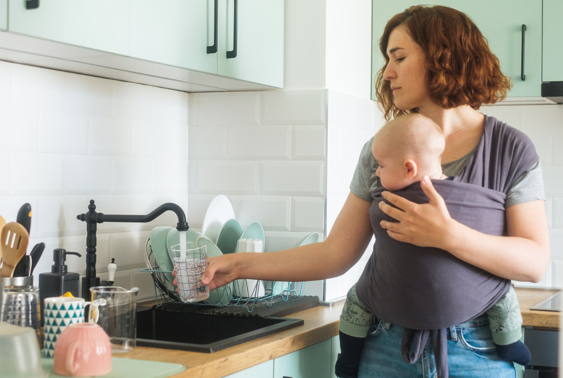 Eine Frau traegt ihr Baby in einem Tragetuch vor dem Bauch und befuellt ein Glas Wasser am Wasserhahn mit einer Hand