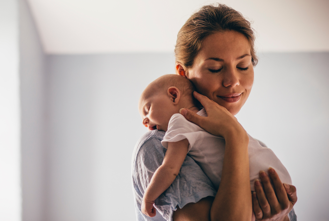 Eine Frau haelt ihr Baby ueber die Schulter und laechelt das Baby gaehnt