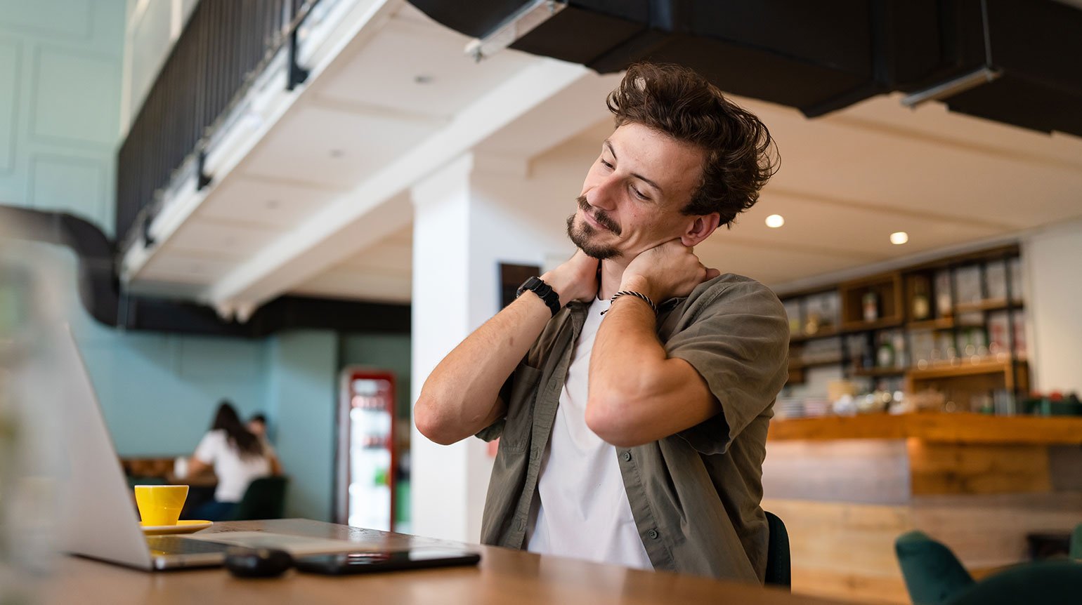 Mann sitzt in einem Café am Tisch, vor sich Laptop, Handy, Tasse. Hinter ihm ein Tresen und zwei weitere Personen im Gespräch. Er legt den Kopf schief und fasst sich mit den Händen rechts und links an die Halswirbelsäule.