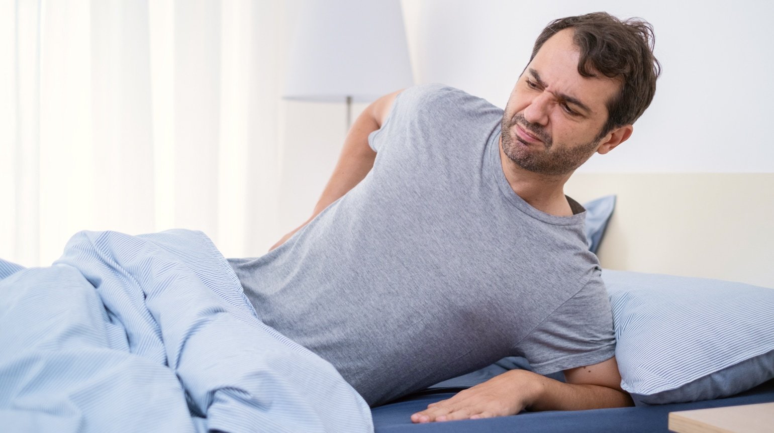 Ein dunkelhaariger Mann liegt auf der Seite im Bett, wobei er sich mit einem Arm abstuezt und mit dem anderen sich vor Schmerzen an der unteren Ruecken fasst