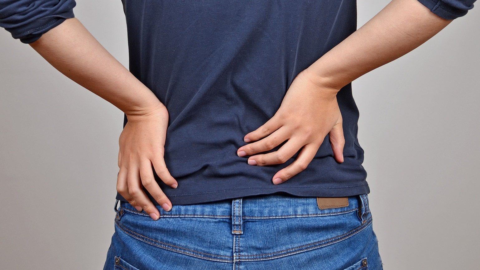 Eine Person in Jeans und T-Shirt hält sich vor Schmerzen den Rücken mit beiden Händen