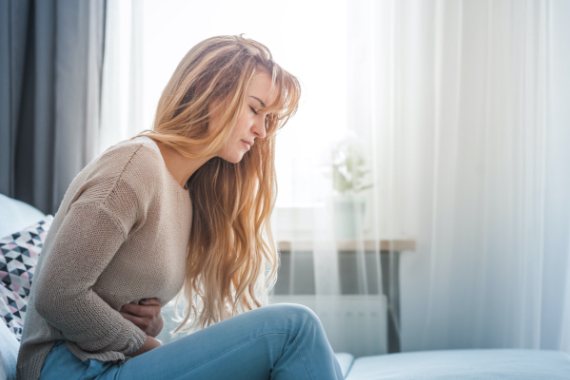 Eine blonde Frau sitzt auf ihrem Bett und hält sich den schmerzenden, verkrampften Unterleib Eine blonde Frau sitzt auf ihrem Bett und hält sich den schmerzenden, verkrampften Unterleib