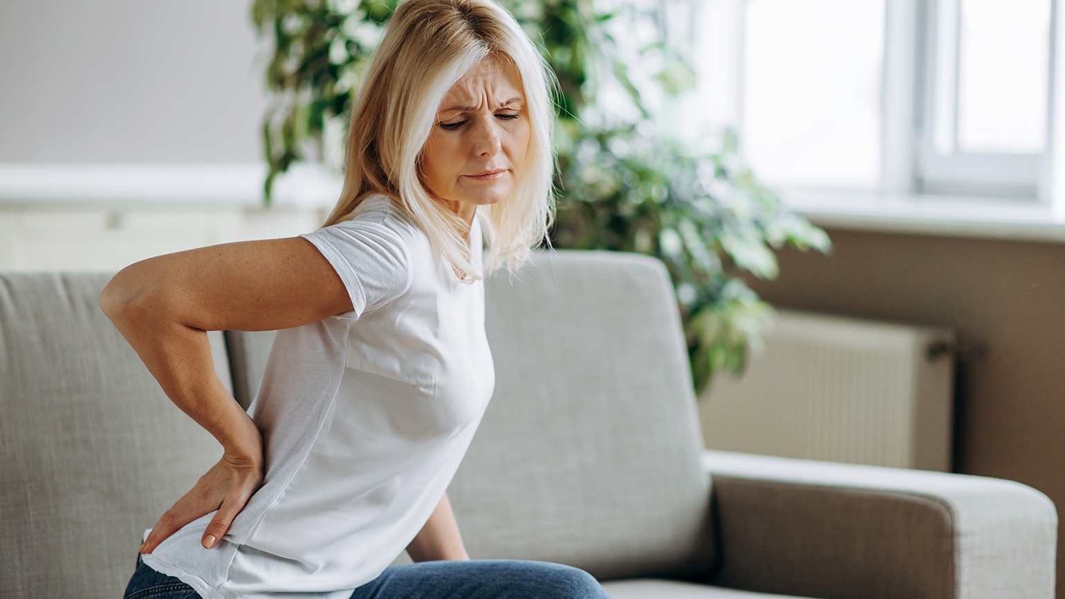 Ältere Frau mit blondem Haar hält sich schmerzhaft den unteren Rücken, während sie auf einem Sofa sitzt. Im Hintergrund sieht man helle Wohnzimmereinrichtung mit Pflanzen.