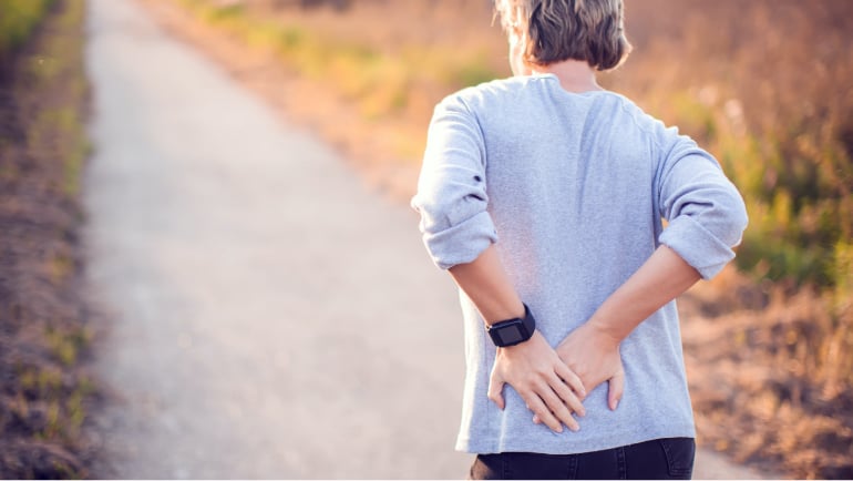 Eine Person geht auf einem Feldweg und haelt sich mit beiden Haenden den unteren Ruecken