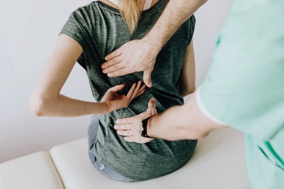 Frau sitzt auf einer Liege sie fasst sich mit einer Hand in den Ruecken ein Mann steht hinter ihr und sie mit beiden Haenden an