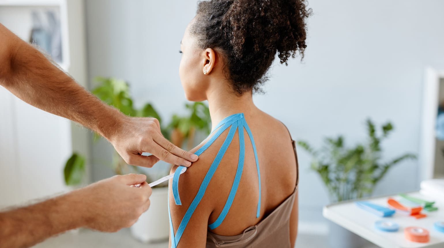 Seitliche Rückenansicht einer sitzenden Frau. Hände eines Physiotherapeuten bringen Kinesio-Taping auf die linke Schulter,  Arm und oberem Rücken an.