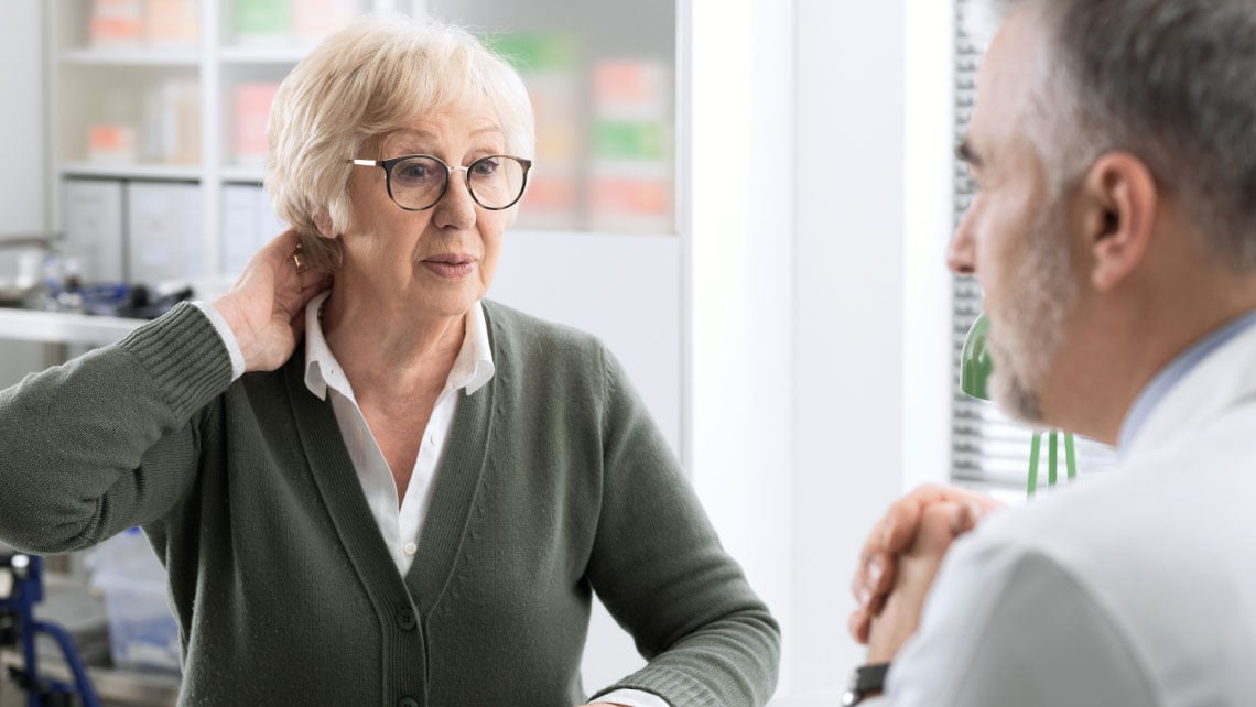 Eine ältere Frau mit Brille sitzt ihrem Arzt gegenüber und beschreibt ihre Nackenschmerzen