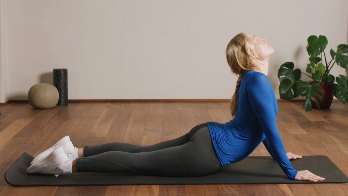 Eine Frau liegt mit dem Bauch nach unten auf einer Gymnastikmatte. Ihre Beine liegen auf, ihre Arme sind durchgestreckt und stützen den Oberkörper nach oben. Den Kopf legt sie in den Nacken.